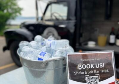Shake & Stir Bar Truck Book Us sign with QR code beside a bucket of chilled water bottles, vintage truck in the background, Irvington NY